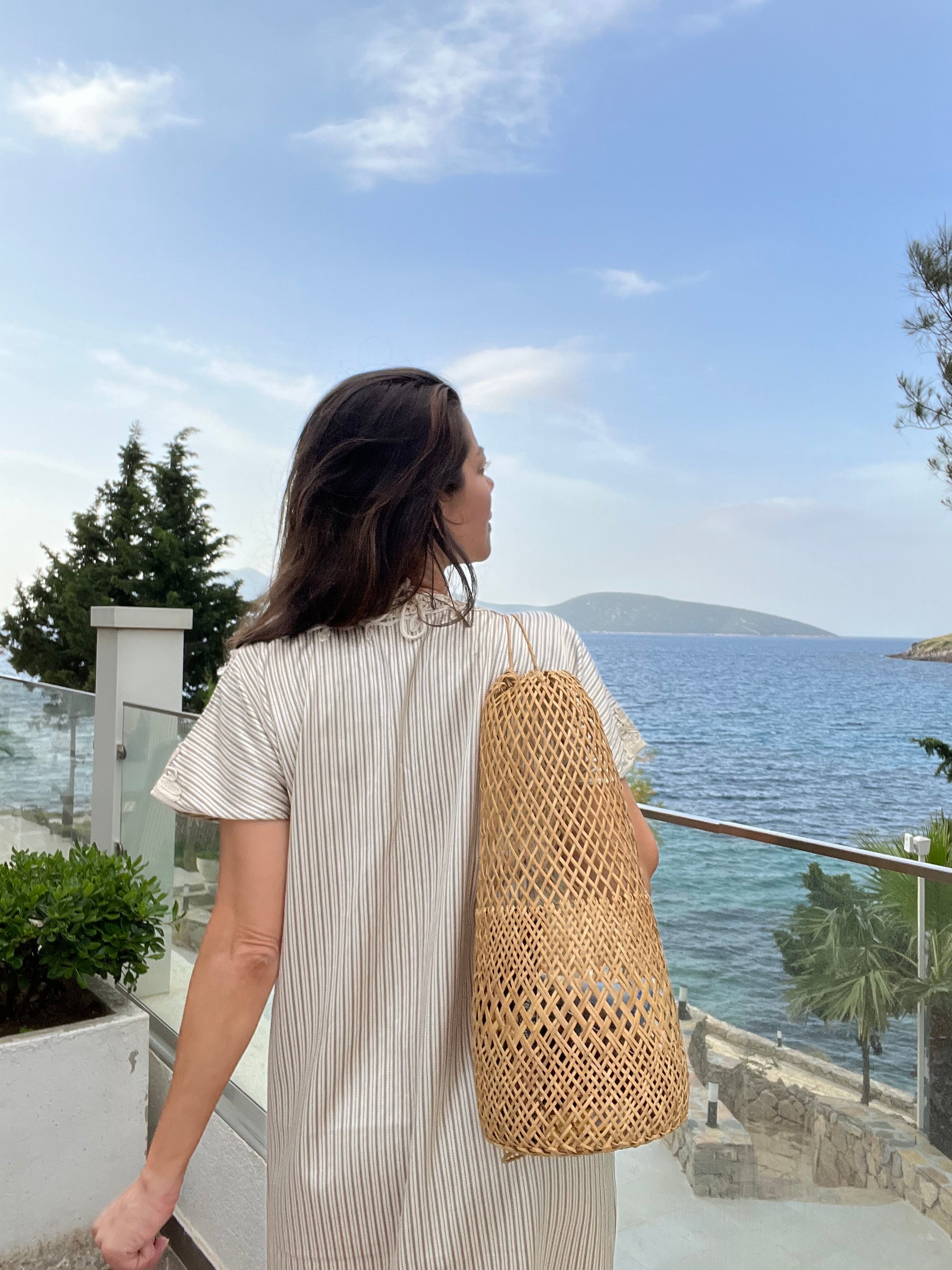 Lifestyle shot of the rattan basket bag worn by a model in a striped linen dress, overlooking a serene Mediterranean coastal view.