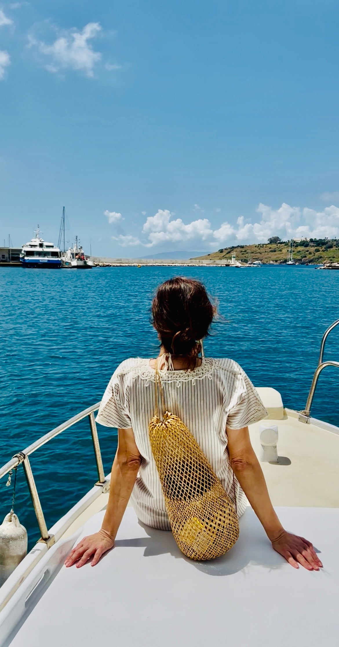 Rear view of the handwoven rattan bag styled as a crossbody on a boat, set against clear blue ocean waters and a summer horizon.