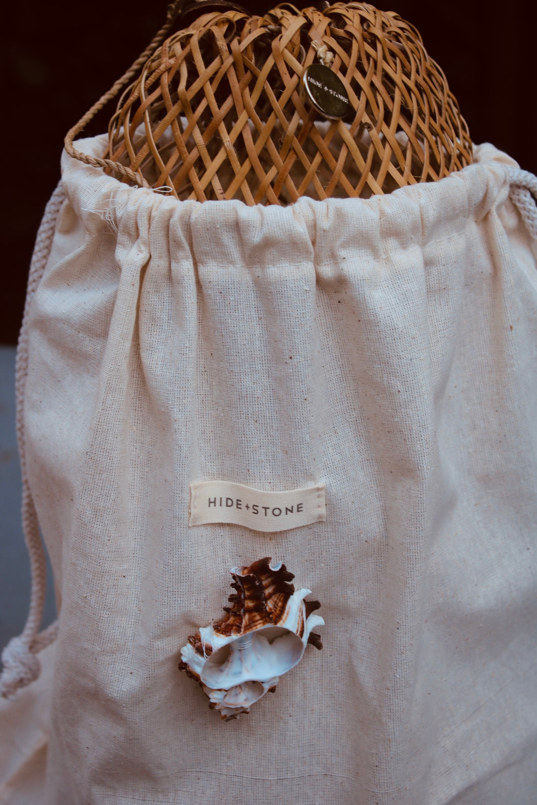 Close-up of the organic cotton dust bag with the Hide + Stone logo and a murex shell detail, tucked inside the rattan weave.