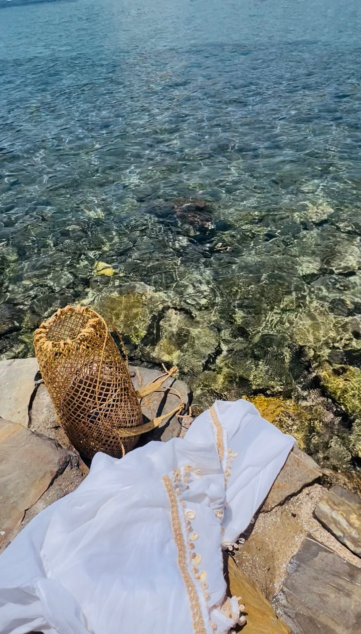 The artisanal rattan bag resting on coastal rocks next to a white embroidered beach kaftan by the turquoise sea.