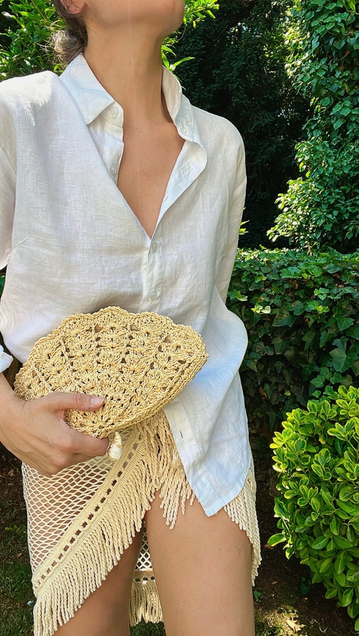A model wearing a white linen shirt and a crocheted fringed skirt holds the handcrafted raffia shell clutch in a garden setting.