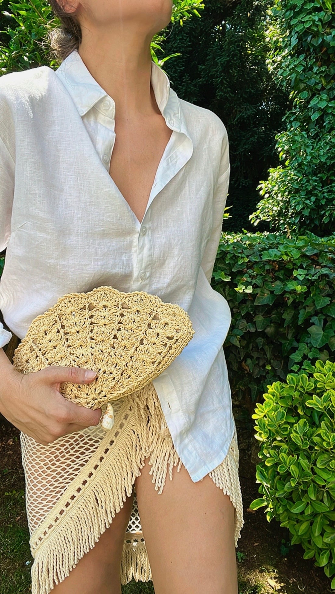 A model wearing a white linen shirt and a crocheted fringed skirt holds the handcrafted raffia shell clutch in a garden setting.