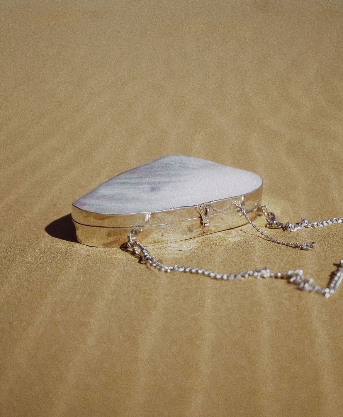 Handcrafted silver-framed hard-shell clutch with an iridescent river mussel shell surface, resting on golden sand with a chain strap attached.