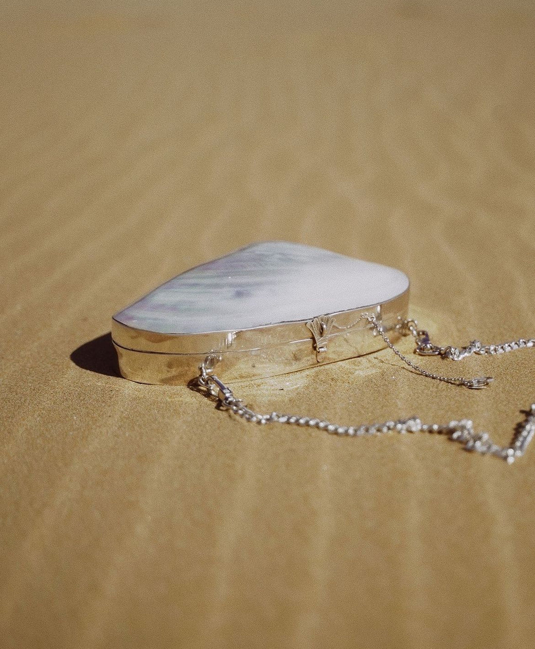 Handcrafted silver-framed hard-shell clutch with an iridescent river mussel shell surface, resting on golden sand with a chain strap attached.