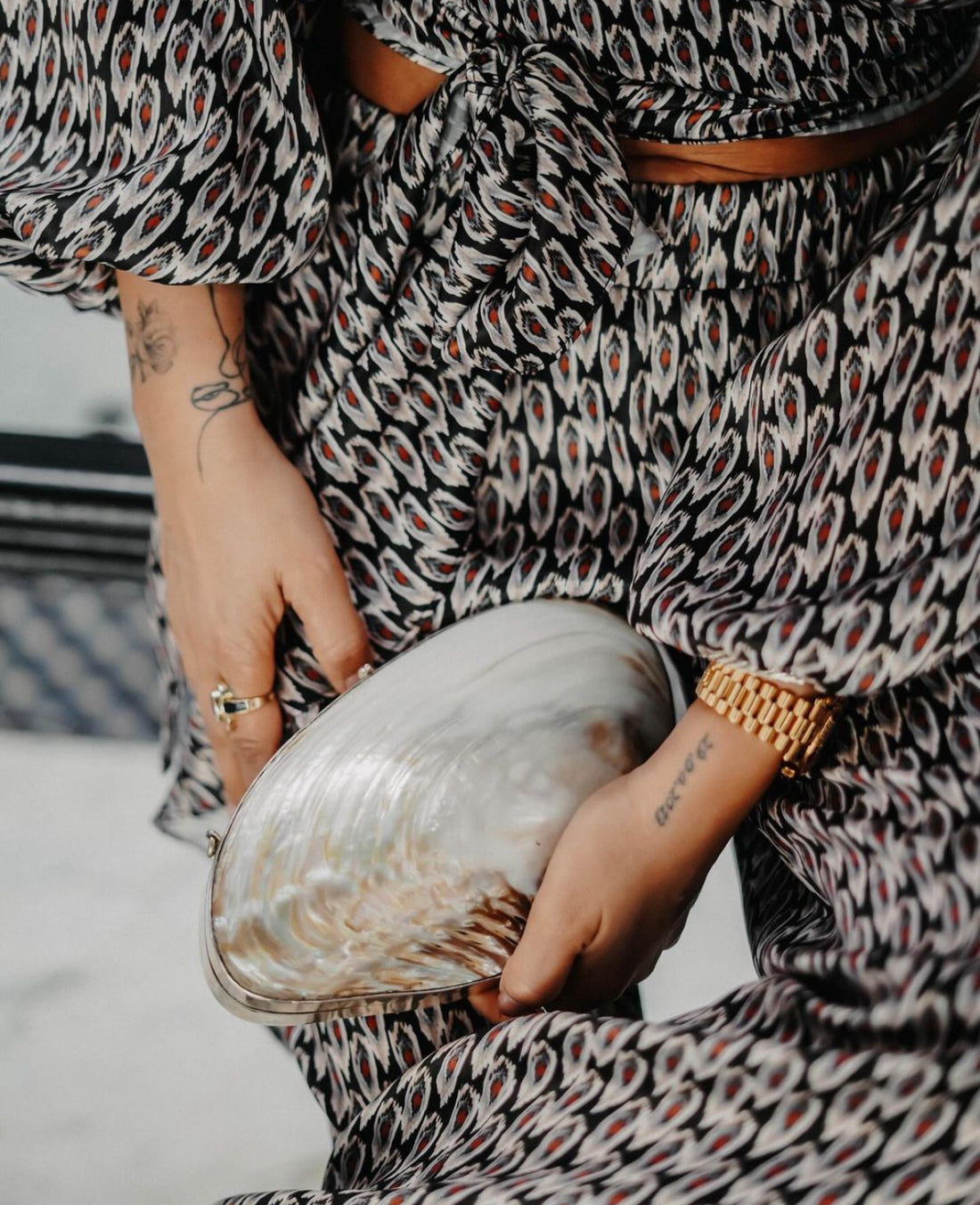 A close-up view of a person wearing a patterned top and a gold watch, holding the handcrafted, silver-framed river mussel shell clutch. 