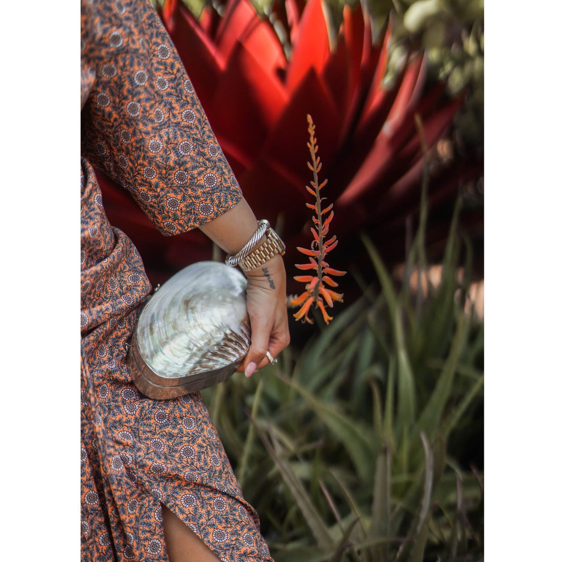 A high-fashion lifestyle shot of a person in an orange floral dress holding the handcrafted silver-framed river mussel shell clutch against a garden background of succulent plants and an aloe flower.