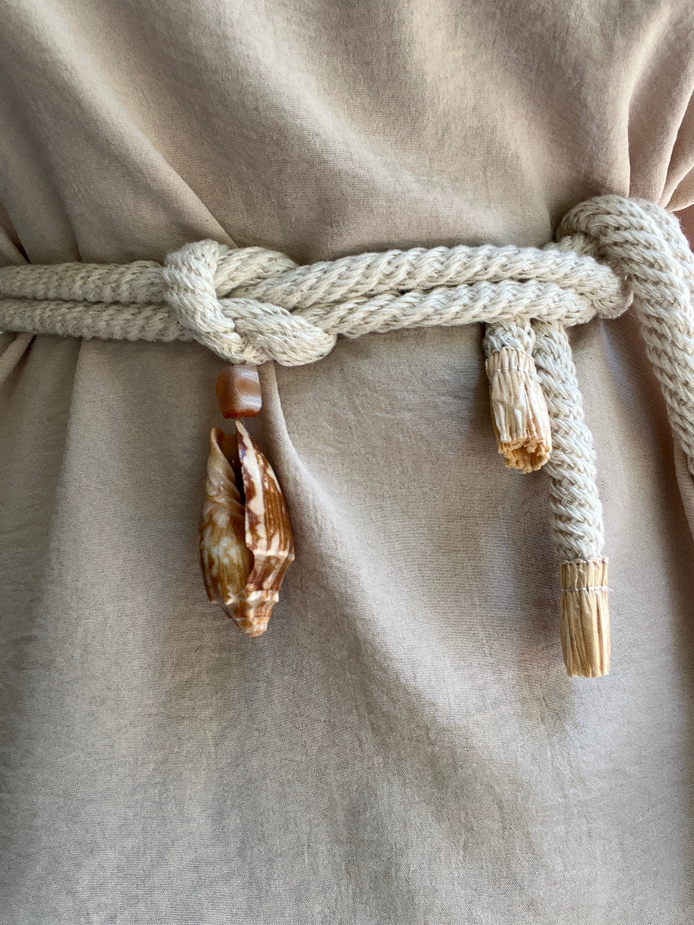 Close-up of a beige linen garment tied with a thick cream cotton rope belt and decorated with cowrie shells and Madagascar quartz.