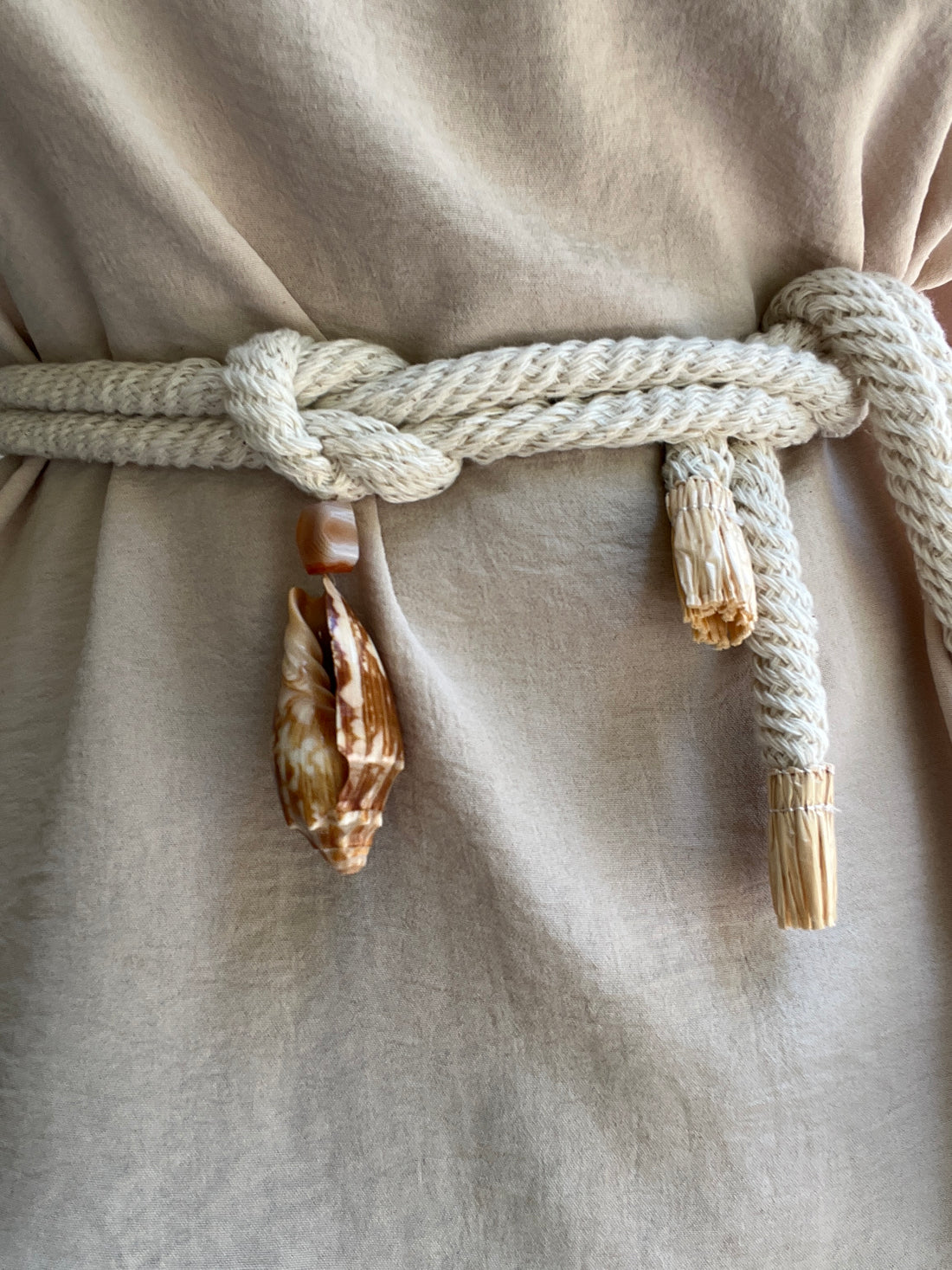 Close-up of a beige linen garment tied with a thick cream cotton rope belt and decorated with cowrie shells and Madagascar quartz.