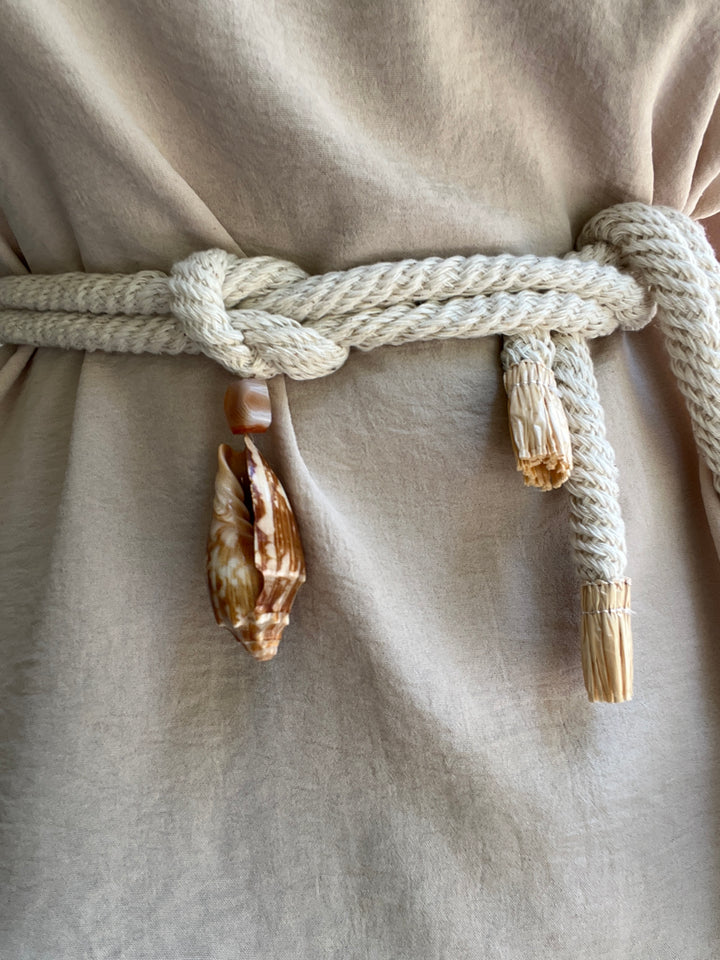 Close-up of a beige linen garment tied with a thick cream cotton rope belt and decorated with cowrie shells and Madagascar quartz.