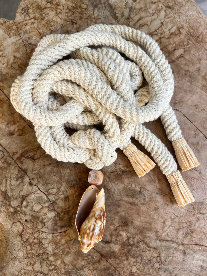 A thick braided organic cotton rope belt with three tassel ends and a cowrie shell pendant and Madagascar quartz, arranged on a textured wood surface.