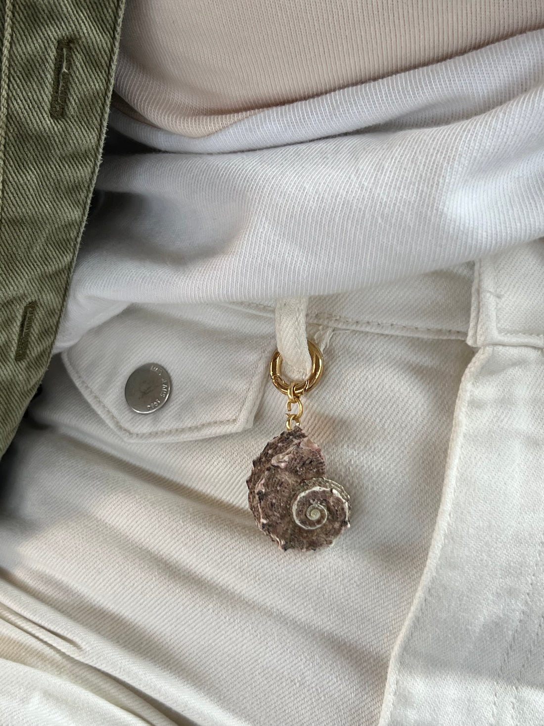 Handcrafted fob featuring a natural, textured spiral shell in earthy purple and grey tones, attached to a gold-toned spring ring styled on off-white jeans