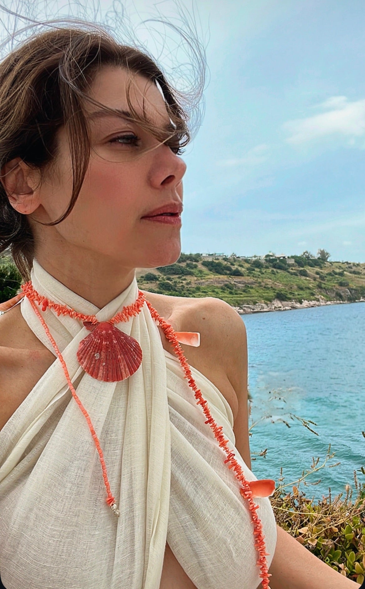 Woman wearing a white top with red coral necklace, standing by a scenic waterfront.