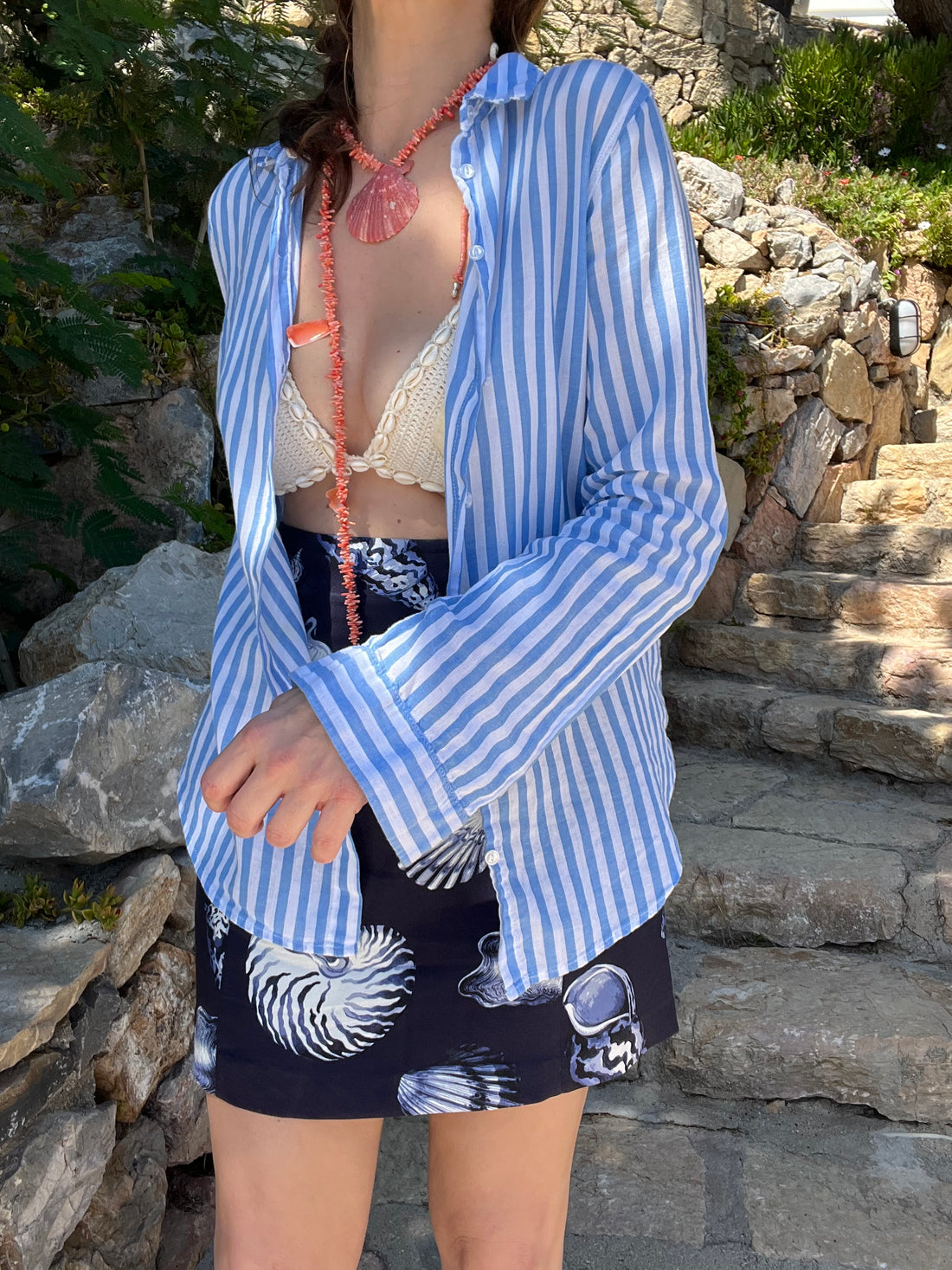 Person wearing a blue and white striped shirt with a black, the red coral necklace  and white crochet swimsuit underneath, standing against a stone wall.