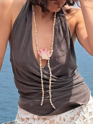 Handcrafted shell necklace featuring a central scallop shell and a strand of tiny shells, finished with white pearls and pink Madagascar rose quartz drops styled on a model on a yacht.