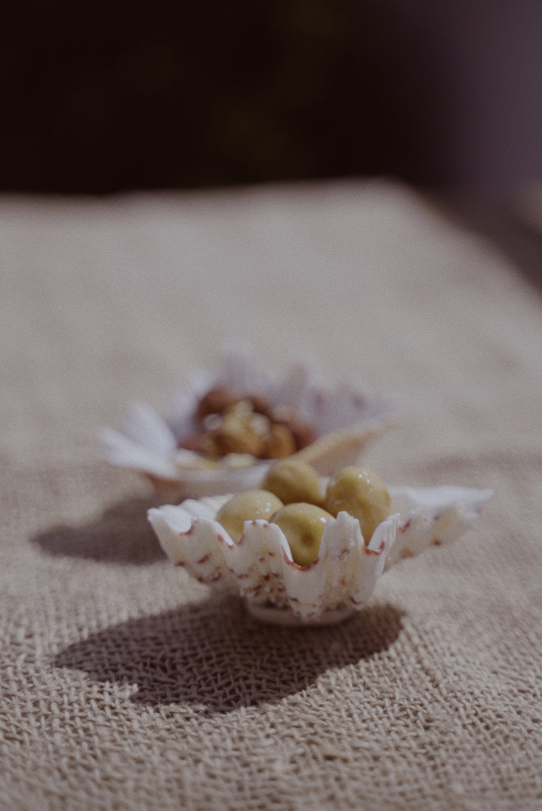 Two white clam shell-shaped serving bowls with a textured surface, displayed on a linen tablecloth. One bowl contains what appears to be olives, and the other has a nut-like snack placed in the center.