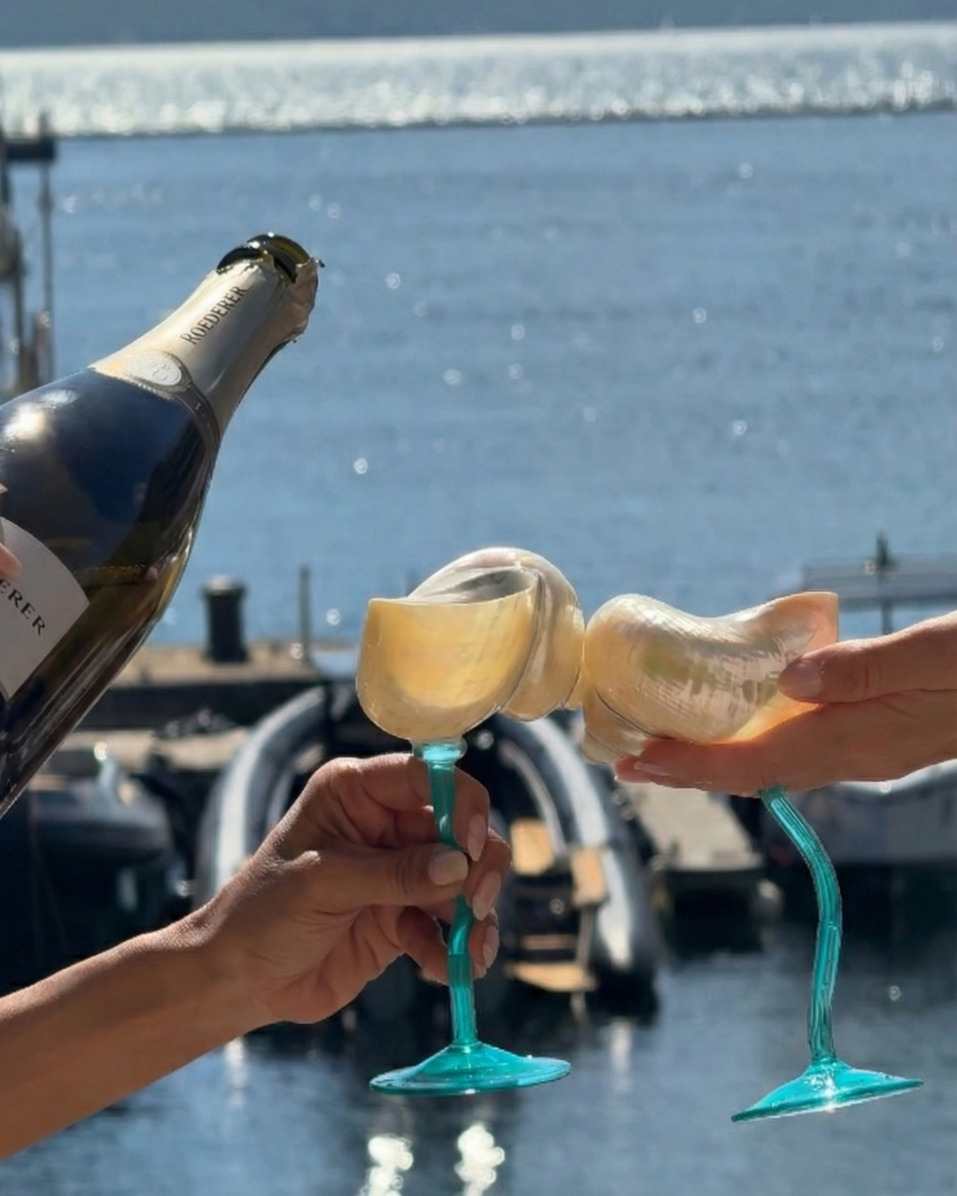 Two people holding the turquoise dream glassware with a bottle of champagne against a waterfront background.