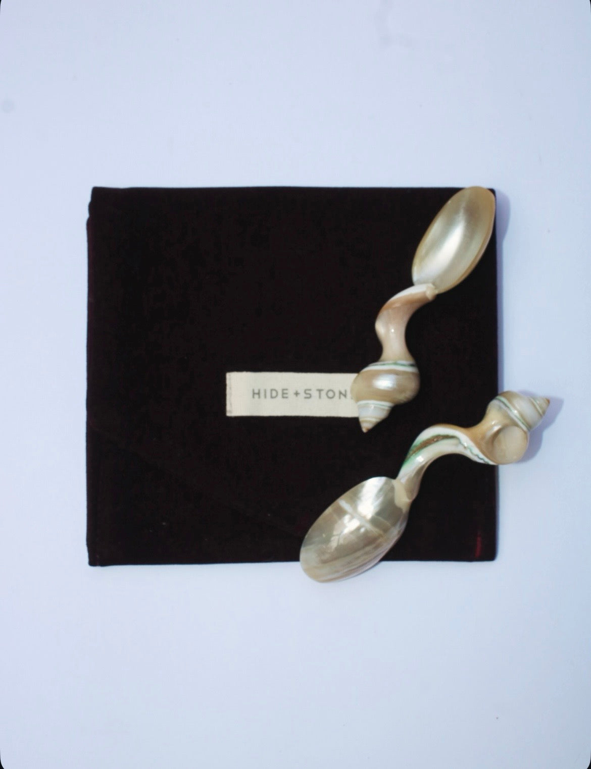 Mother of pearl spoons handcrafted with unique shell  handles on a black pouch with 'HIDE & STONE' branding against a light purple background.