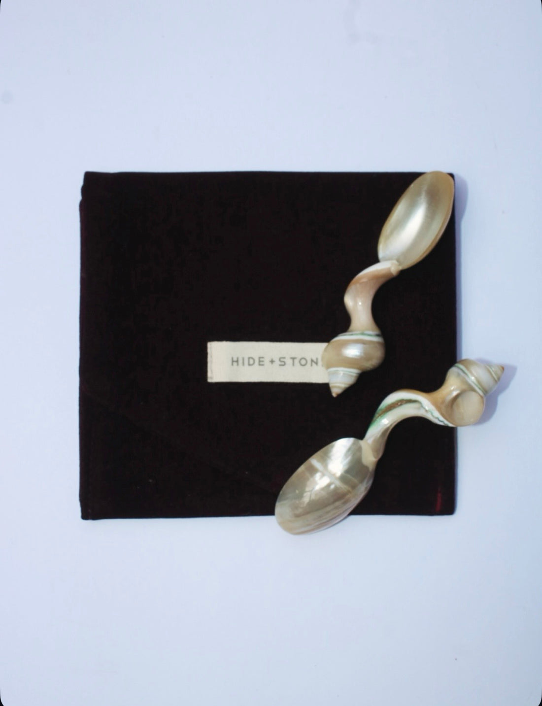 Mother of pearl spoons handcrafted with unique shell  handles on a black pouch with 'HIDE & STONE' branding against a light purple background.