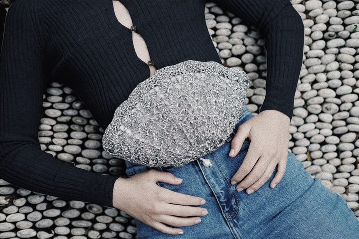 Handcrafted metallic silver crochet shell-shaped clutch held by a model on a textured stone pebble path, featuring a pearl drop.